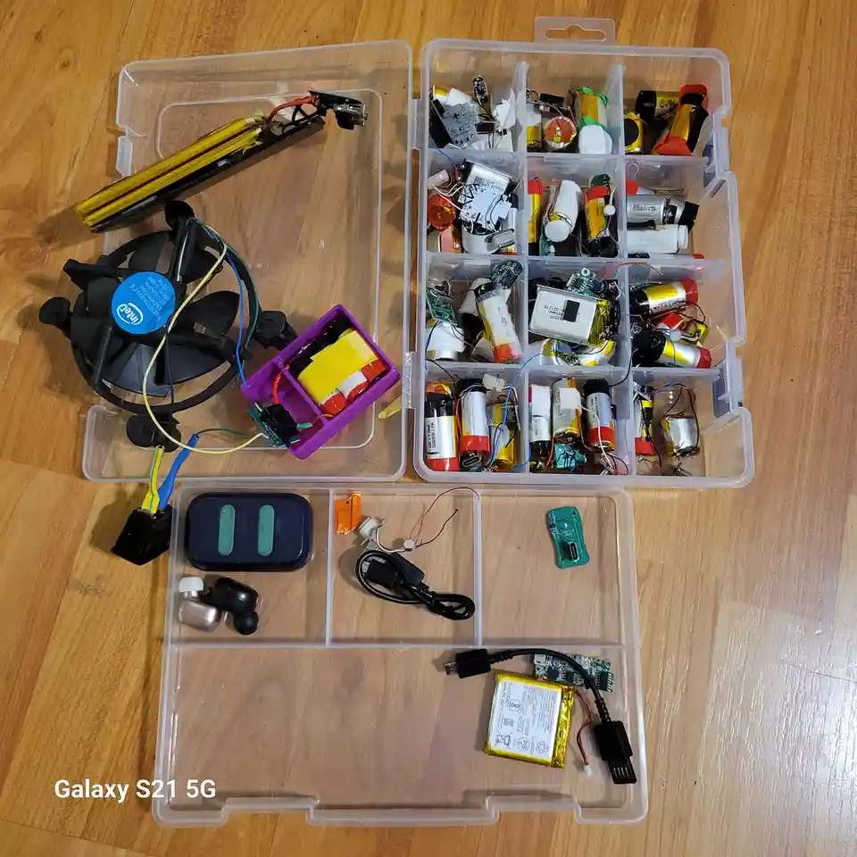Three open plastic storage boxes on a wooden floor contain a disassembled computer fan with an Intel sticker, a purple box…