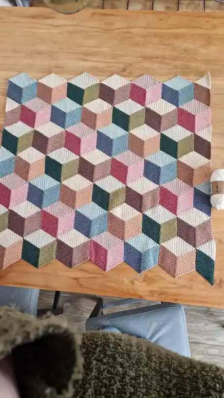 A colorful, hand-knitted blanket with a 3D cube illusion pattern made of multicolored yarn squares is laid out on a wooden…