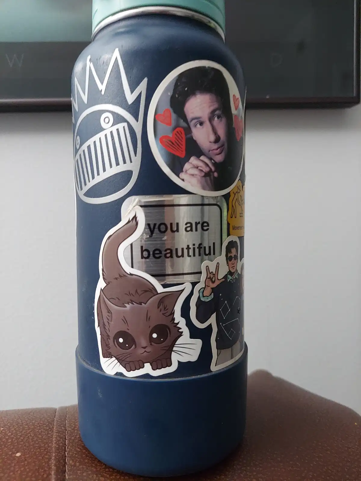 A blue water bottle covered in various stickers, including a circular photo of a man with hearts around him, a cartoon cat…
