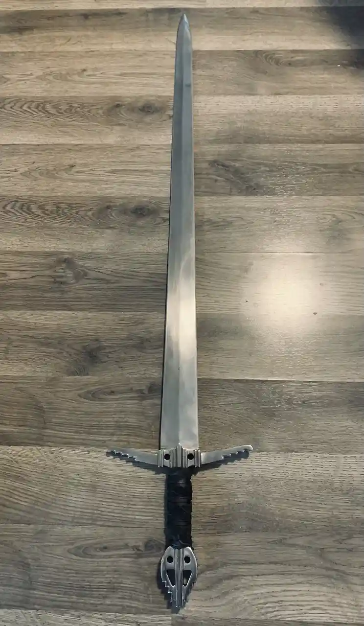 A long, silver-colored sword with a black-wrapped hilt and decorative pommel lies vertically on a gray wood-grain floor. The…