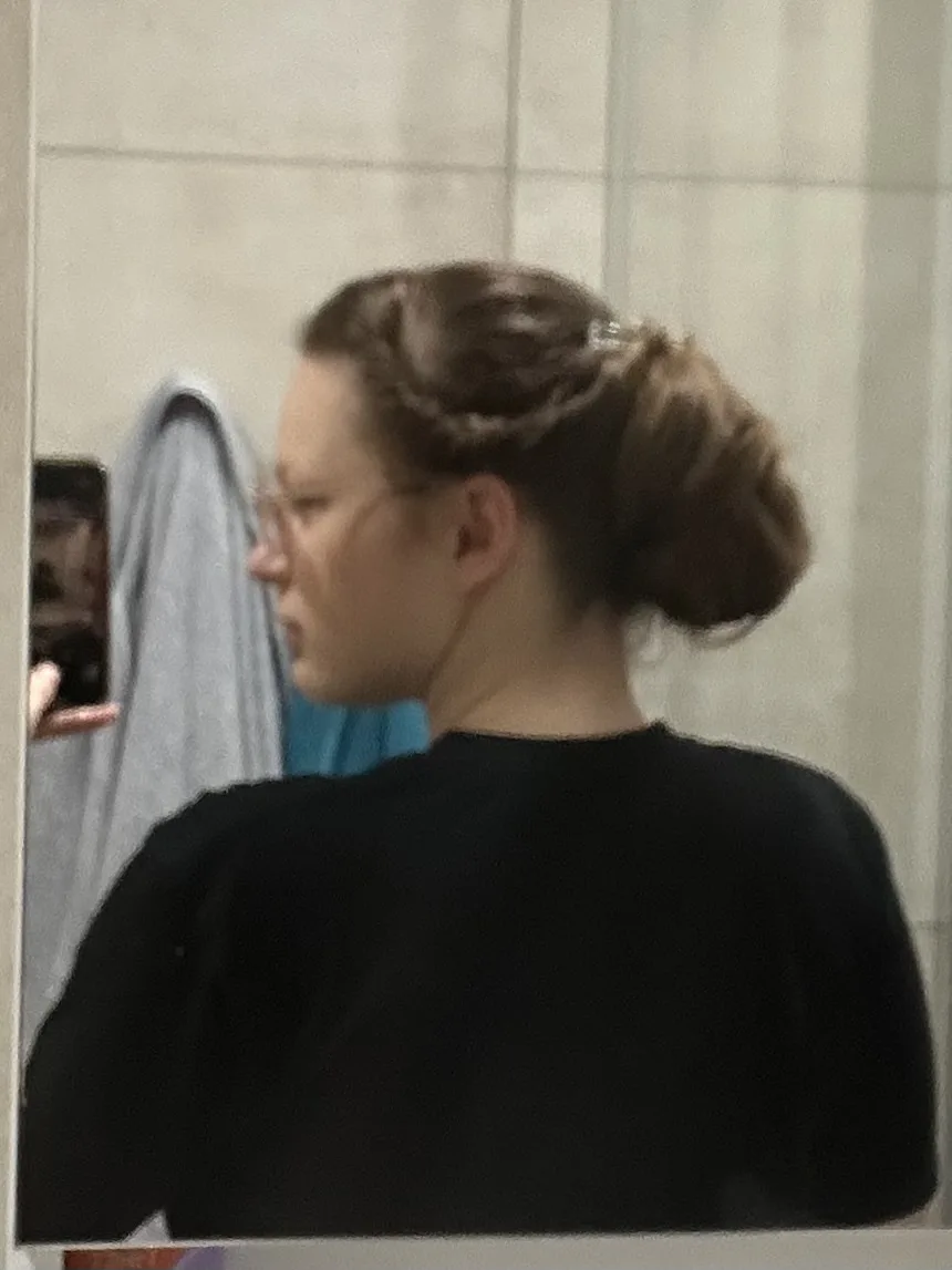 A person with brown hair styled in a messy bun, wearing glasses and a black shirt, takes a mirror selfie in a bathroom. The…