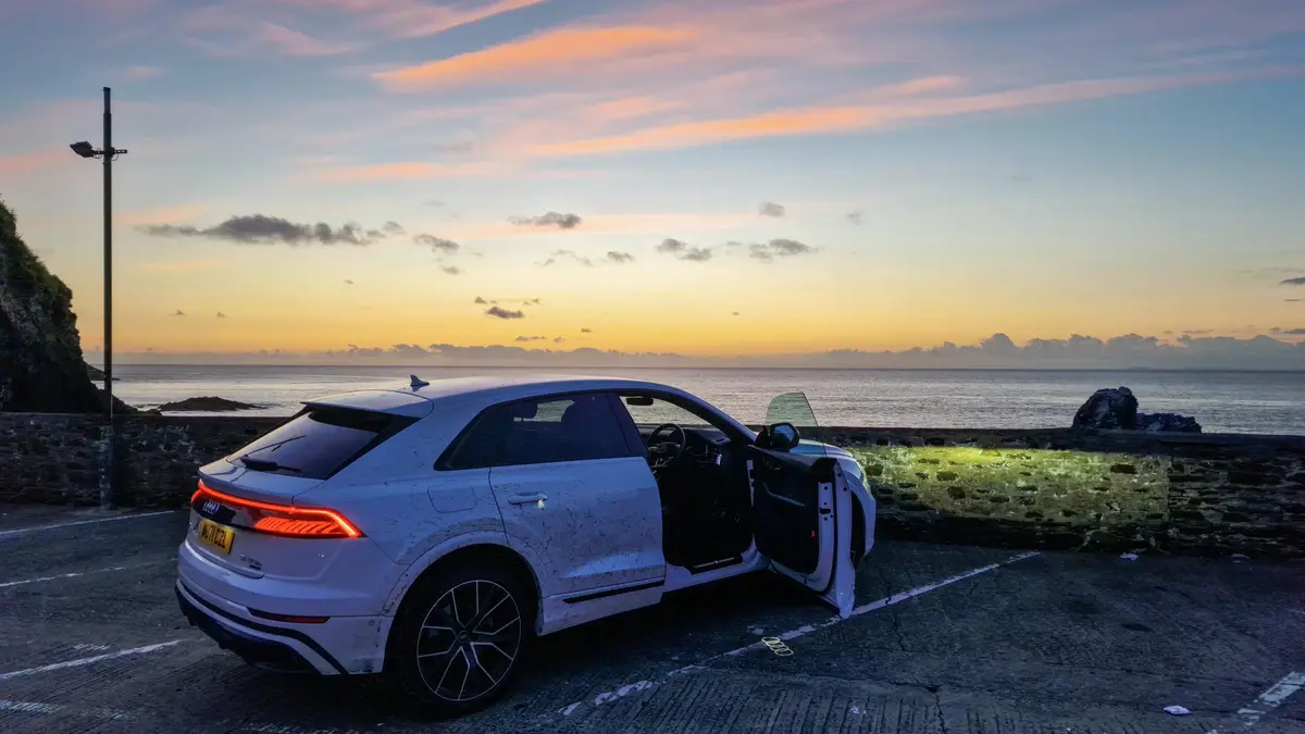 A white Audi SUV is parked in a seaside parking lot at sunset, its driver-side door open, facing the ocean. The car is…