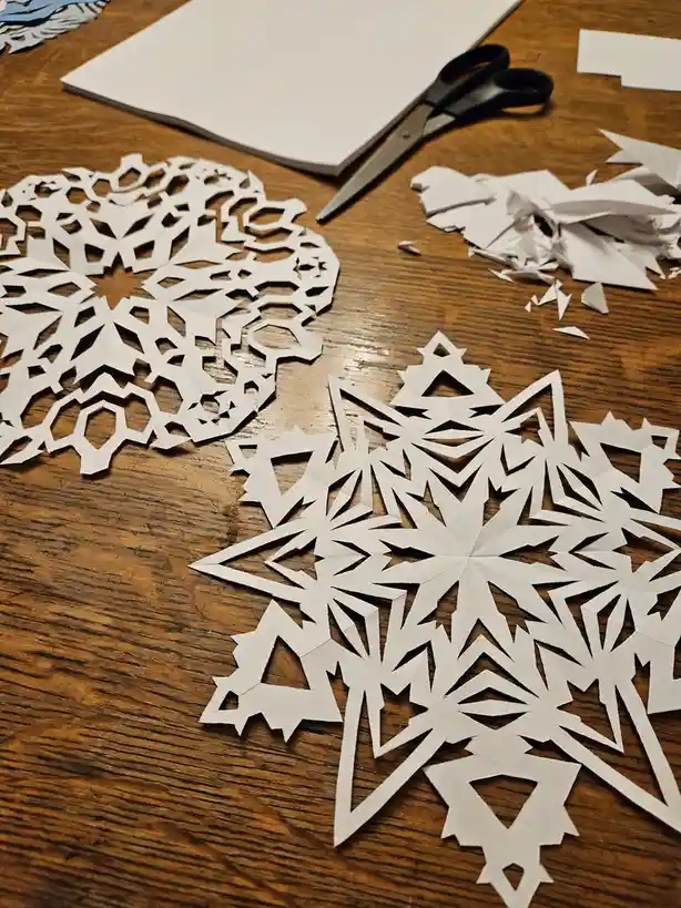A wooden table displays two distinct paper snowflake cutouts, one with a complex geometric pattern and another with a…