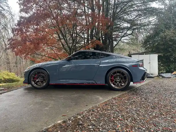 A dark gray Nissan Z sports car is parked on a driveway next to a gravel area, with autumn trees in the background. The car…