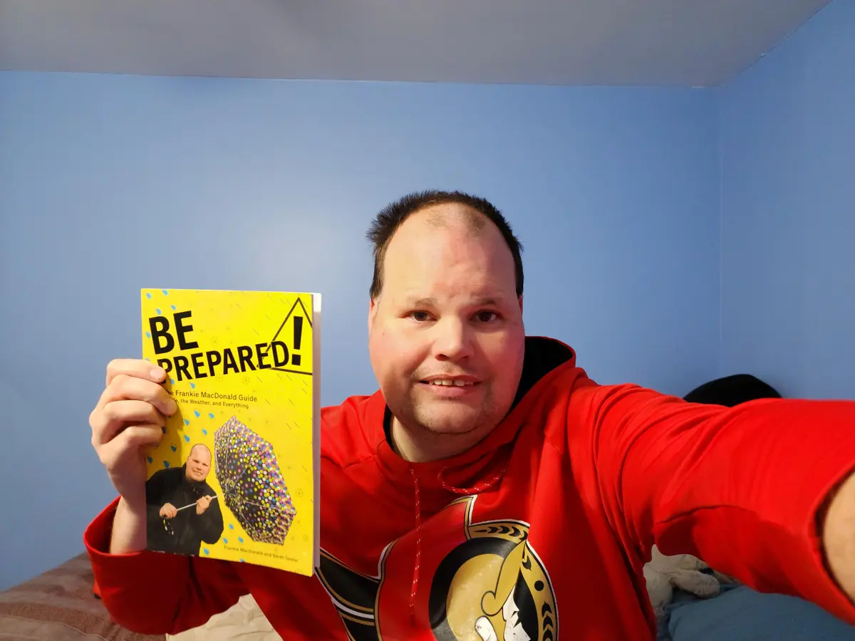 A man in a red hoodie takes a selfie while holding up a bright yellow book titled 'BE PREPARED! The Frankie MacDonald Guide…