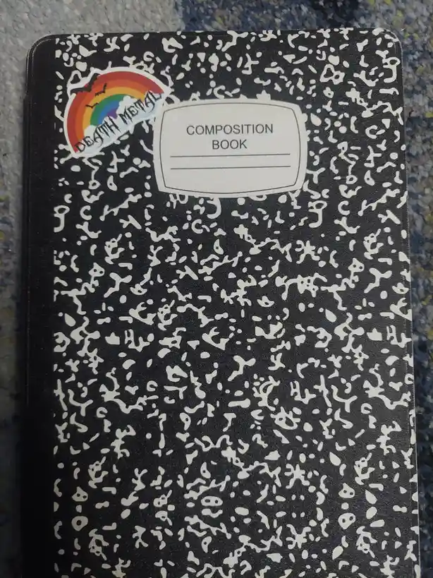 A black composition book with a white speckled pattern has a sticker in the top left corner showing a rainbow with bats…