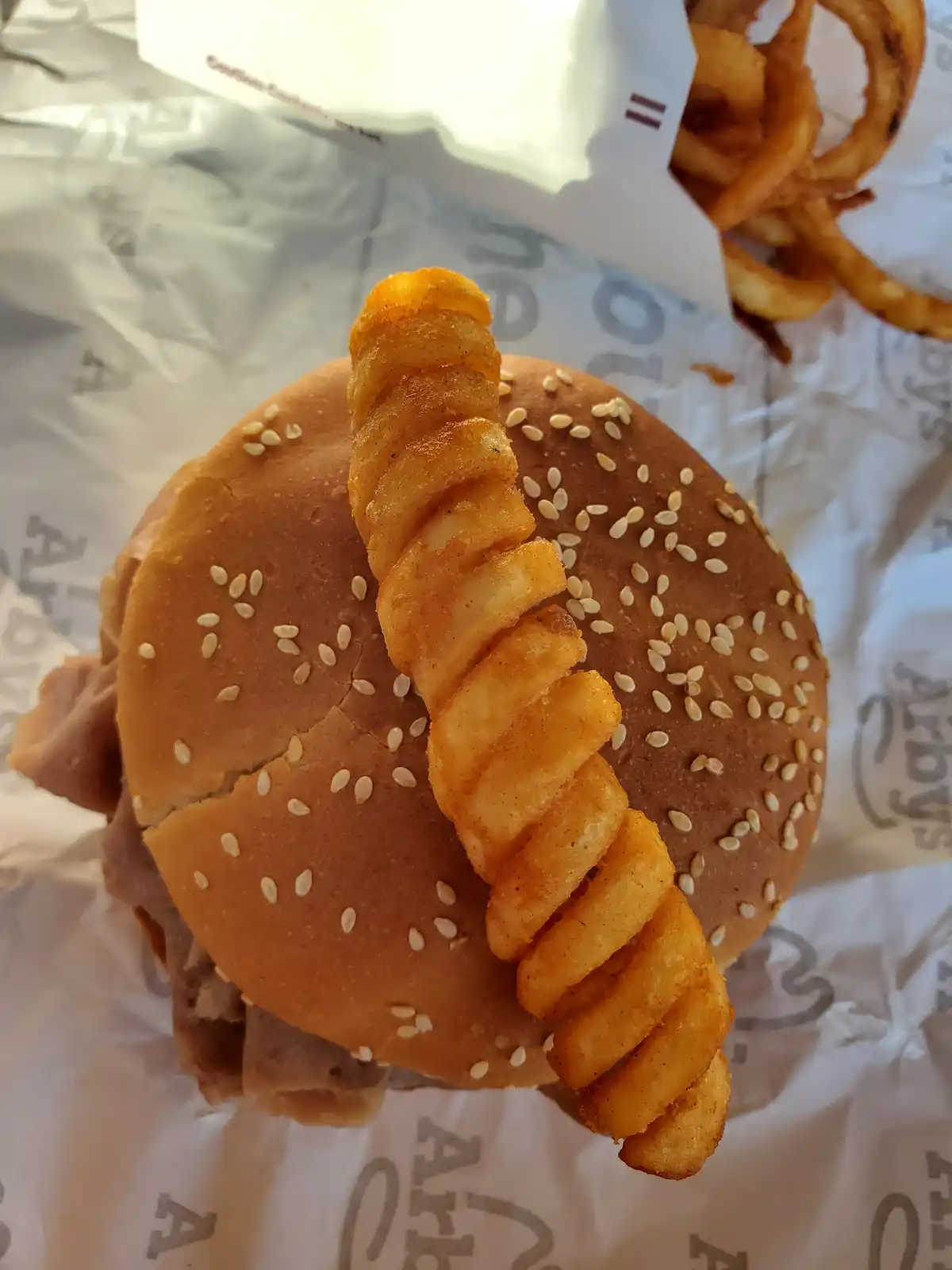 A close-up photo shows a sesame seed burger with a single curly fry stuck vertically through its top bun, resting on Arby's…