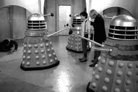 A black-and-white photo shows three Daleks from the British sci-fi series Doctor Who standing in a room. Two Daleks are…