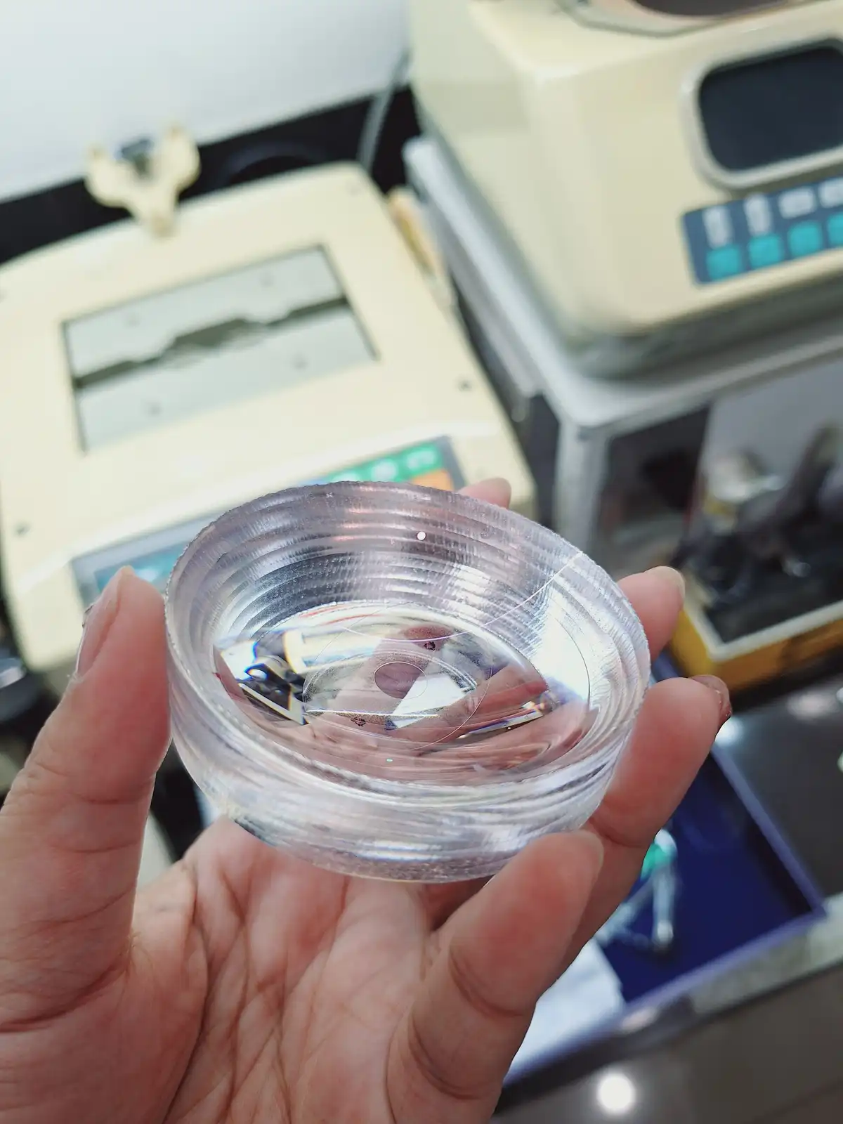 A hand holds a clear, circular, ribbed plastic lens in front of blurred beige lab equipment. The overlaid text reads 'i made…