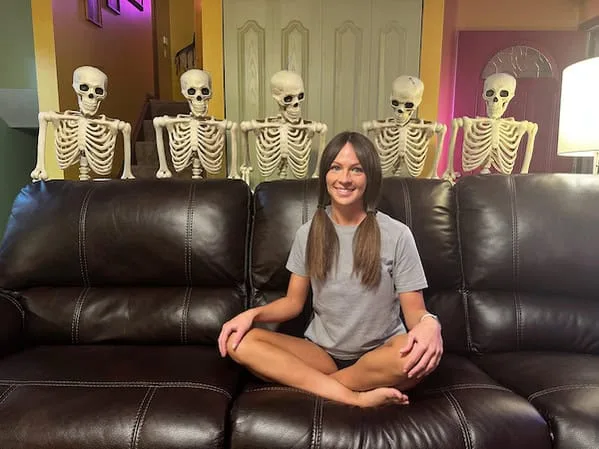 A woman sits cross-legged on a dark brown leather couch, smiling at the camera. Behind her, five plastic skeleton props are…