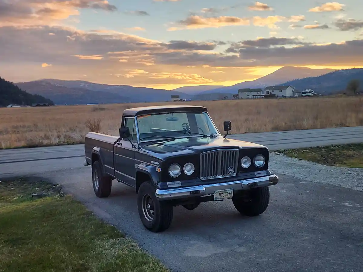 A black vintage Jeep pickup truck is parked on a gravel road at sunset, with mountains and a field in the background. The…