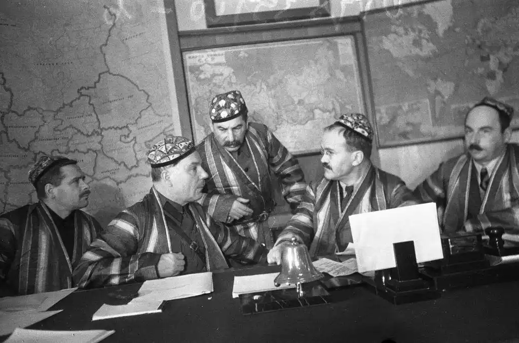 a black-and-white historical photo shows five men in traditional striped robes and patterned hats seated around a conference…