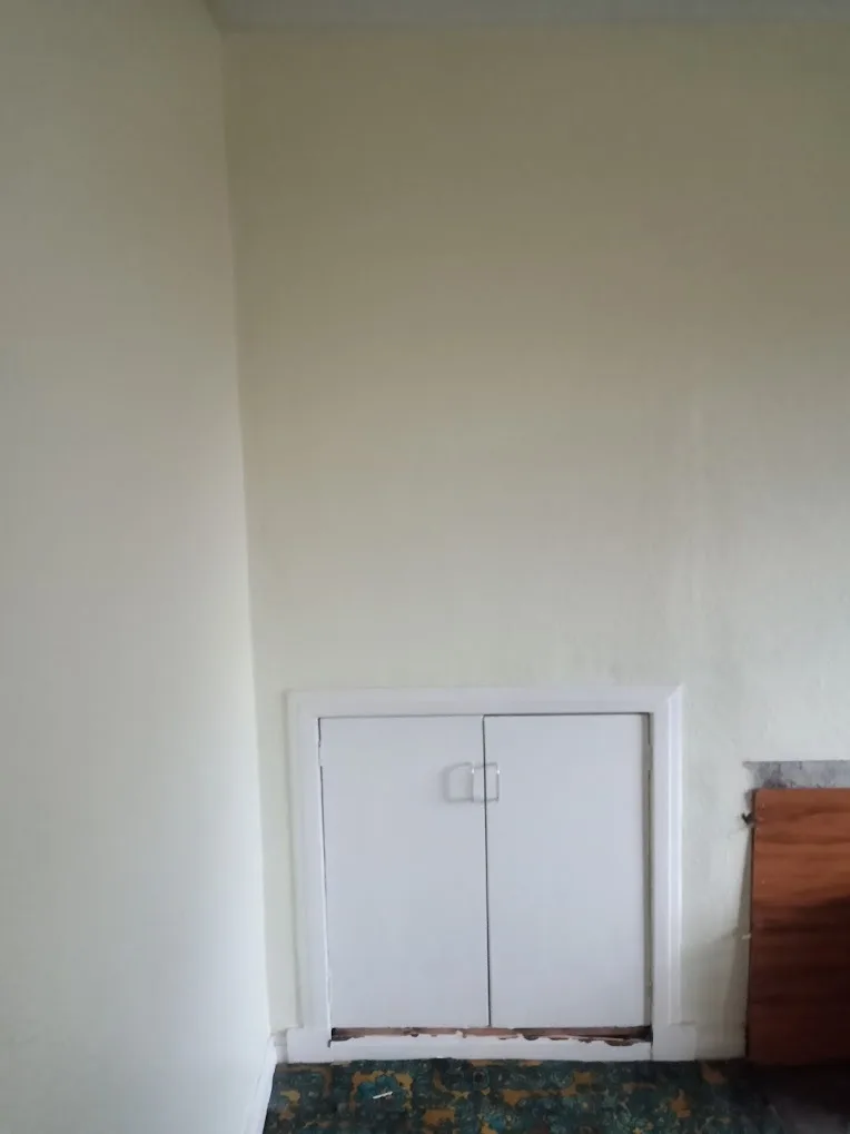 A plain white wall features a small, built-in double-door cabinet with silver handles, set within a white frame. The floor…