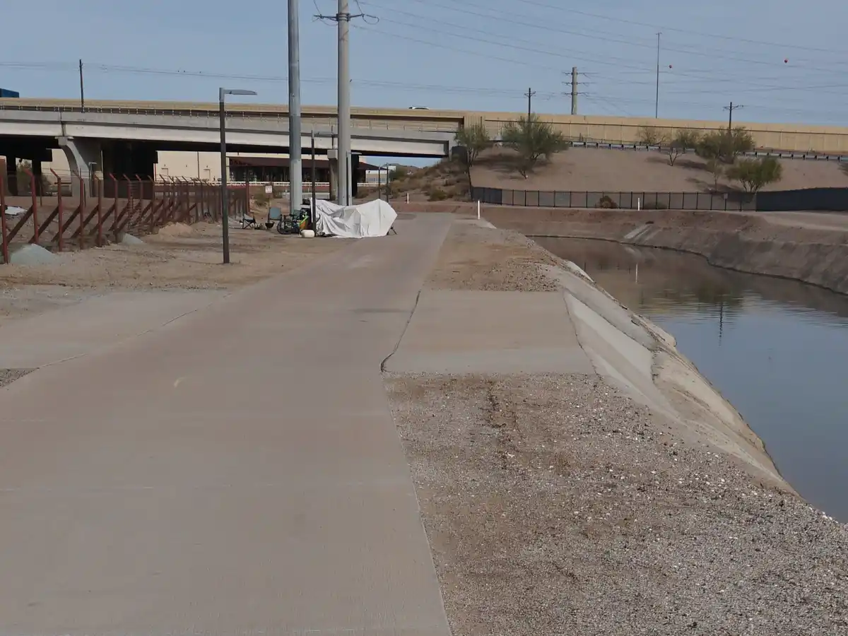 A desolate concrete path runs alongside a canal in a dry, arid landscape under a bridge. A makeshift white tarp shelter with…
