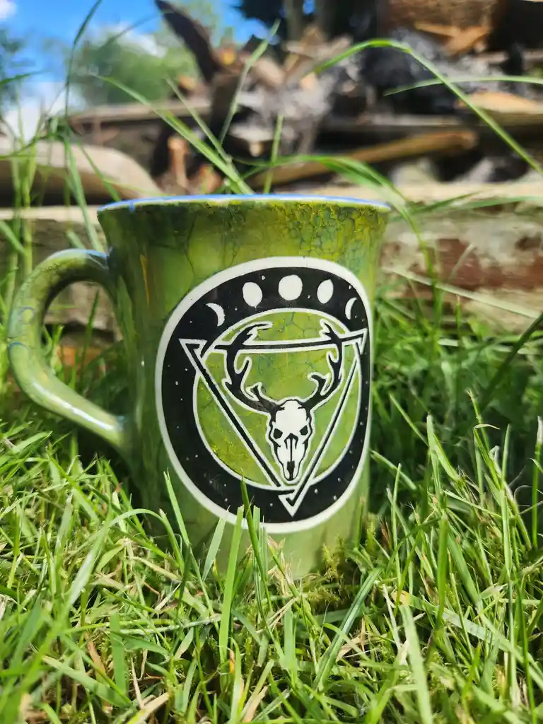A green ceramic mug with a crackled glaze sits in bright green grass, featuring a black-and-white circular emblem of a deer…