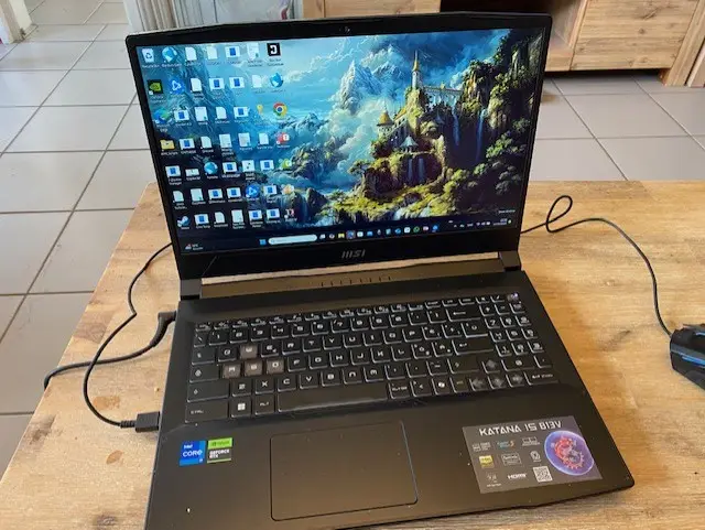 A black MSI Katana 15 laptop sits open on a wooden table, its screen displaying a vibrant fantasy castle wallpaper with many…
