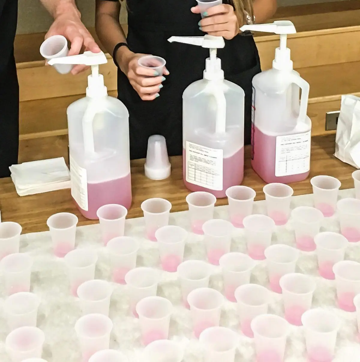 Two people in black shirts are pouring pink liquid from large white pump bottles into small clear plastic cups arranged on a…