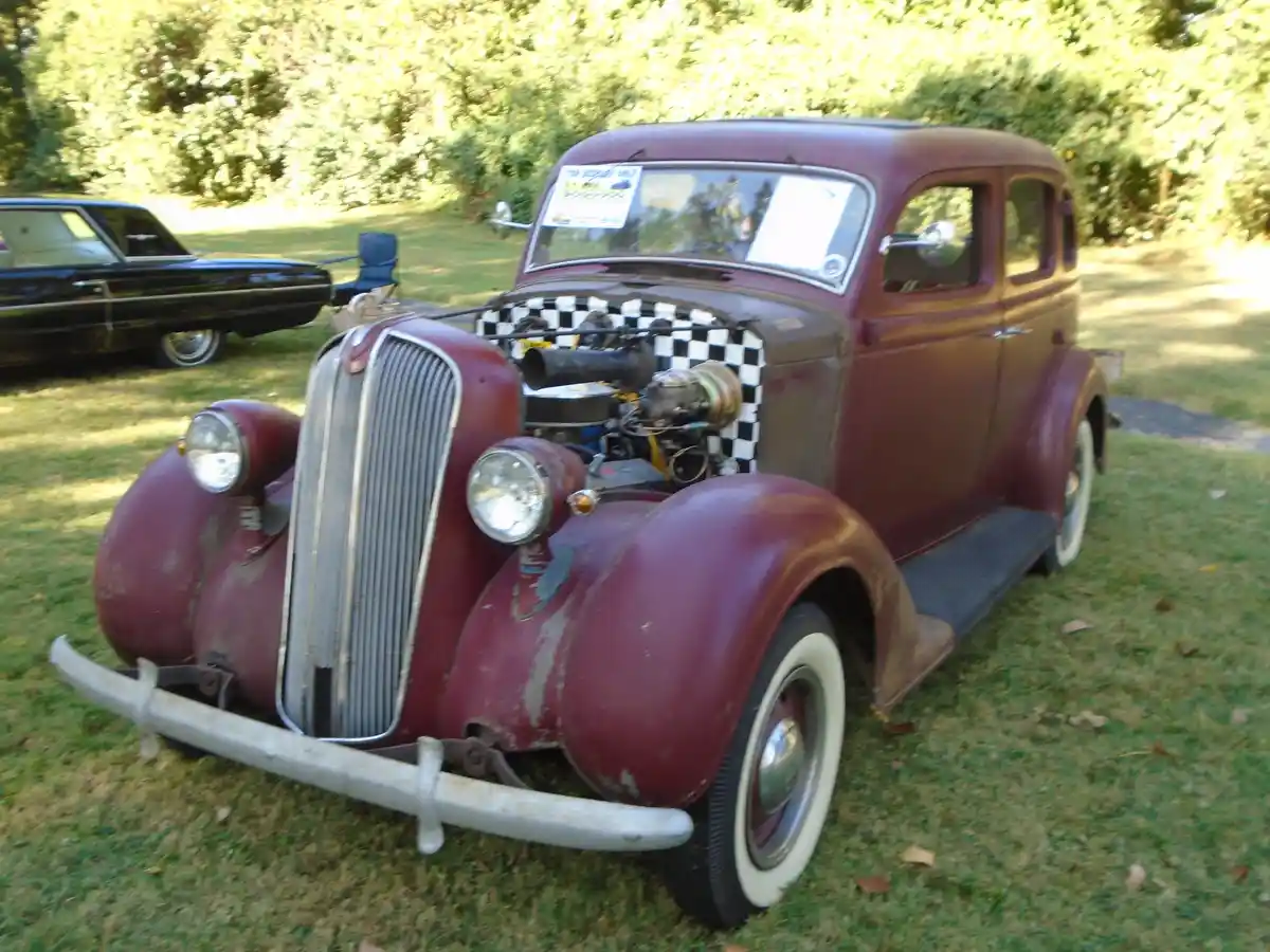 A maroon vintage car with its hood open, revealing a modified engine covered by a black-and-white checkered pattern, parked…