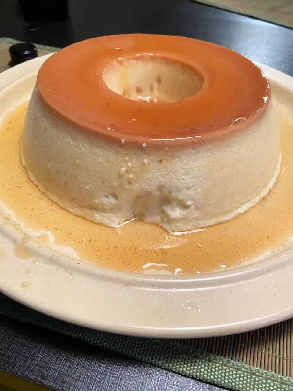 A close-up photo shows a round, creamy flan dessert with a glossy caramel topping, sitting on a white plate. The flan has a…