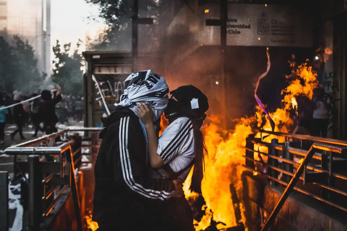 Two people wearing Adidas tracksuits and head coverings embrace passionately in front of a large fire during what appears to…
