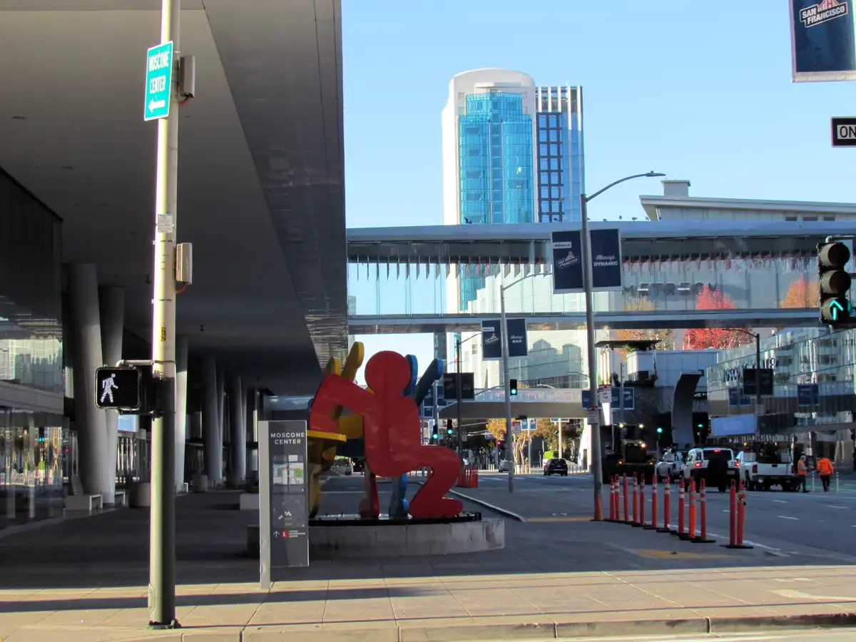 A daytime photo of the Moscone Center in San Francisco, featuring a large colorful abstract sculpture in the foreground, a…