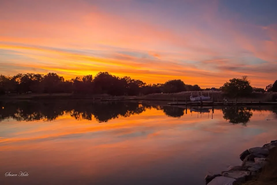 A serene sunset over a calm lake with vibrant orange and pink clouds reflected in the water, trees lining the shore, and a…