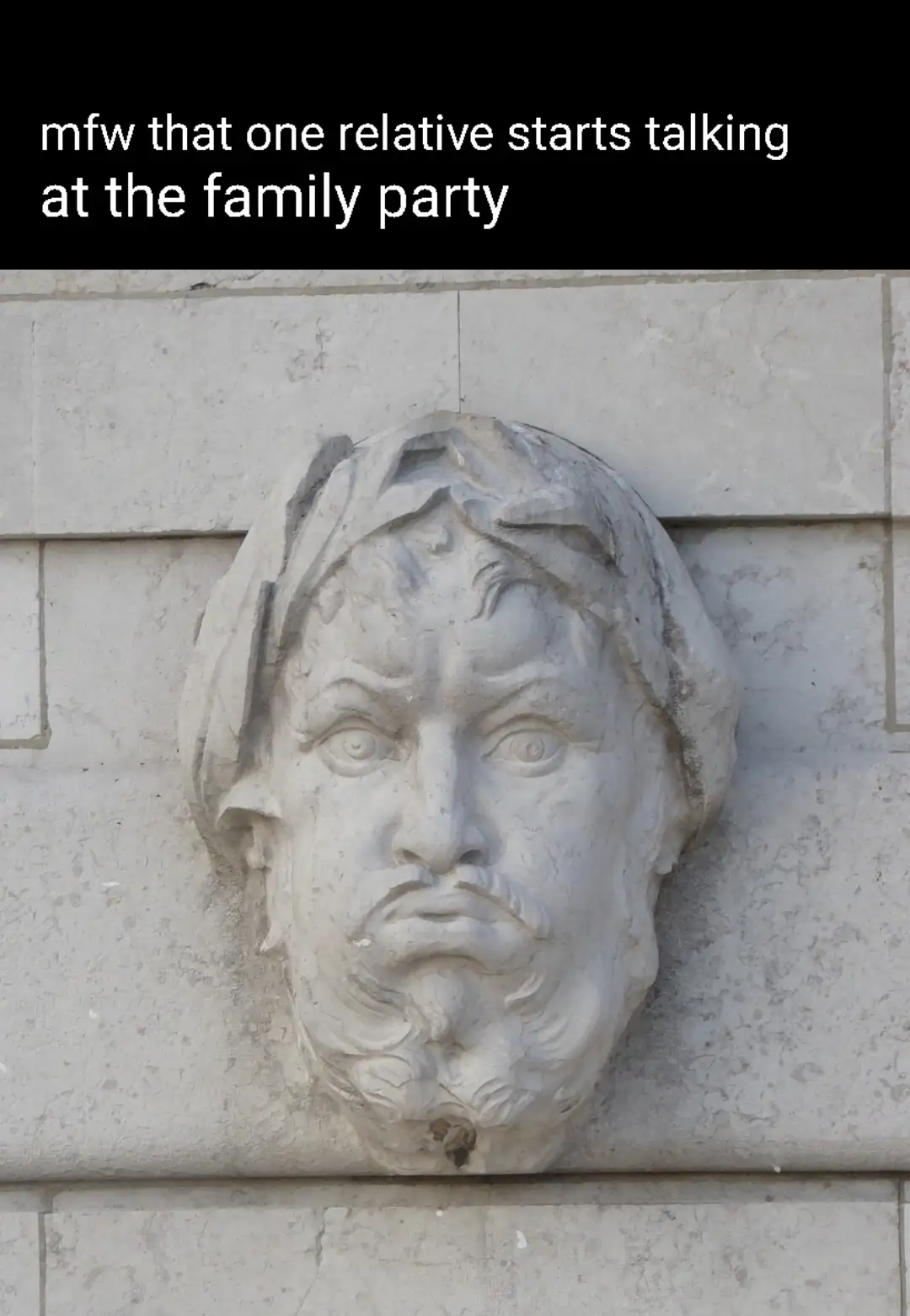 A stone relief sculpture of a man's face with a shocked, wide-eyed, and slightly open-mouthed expression is mounted on a…