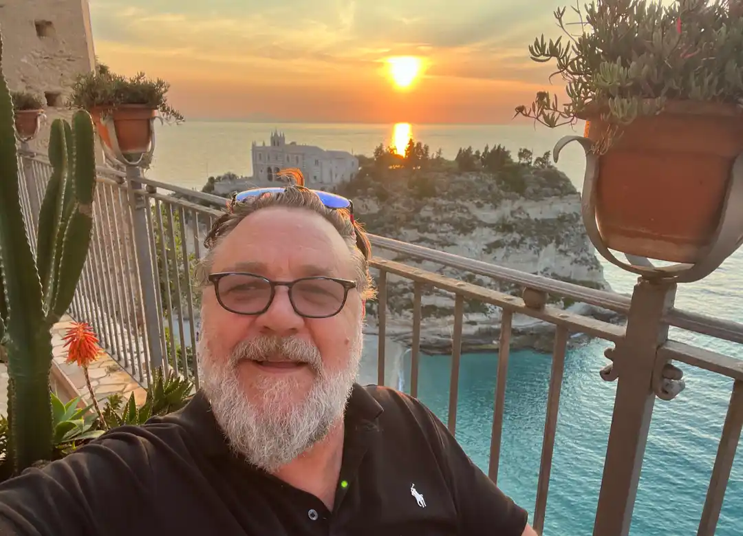 a man with glasses and a white beard takes a selfie on a balcony overlooking a scenic sunset over the ocean, with cliffs, a…