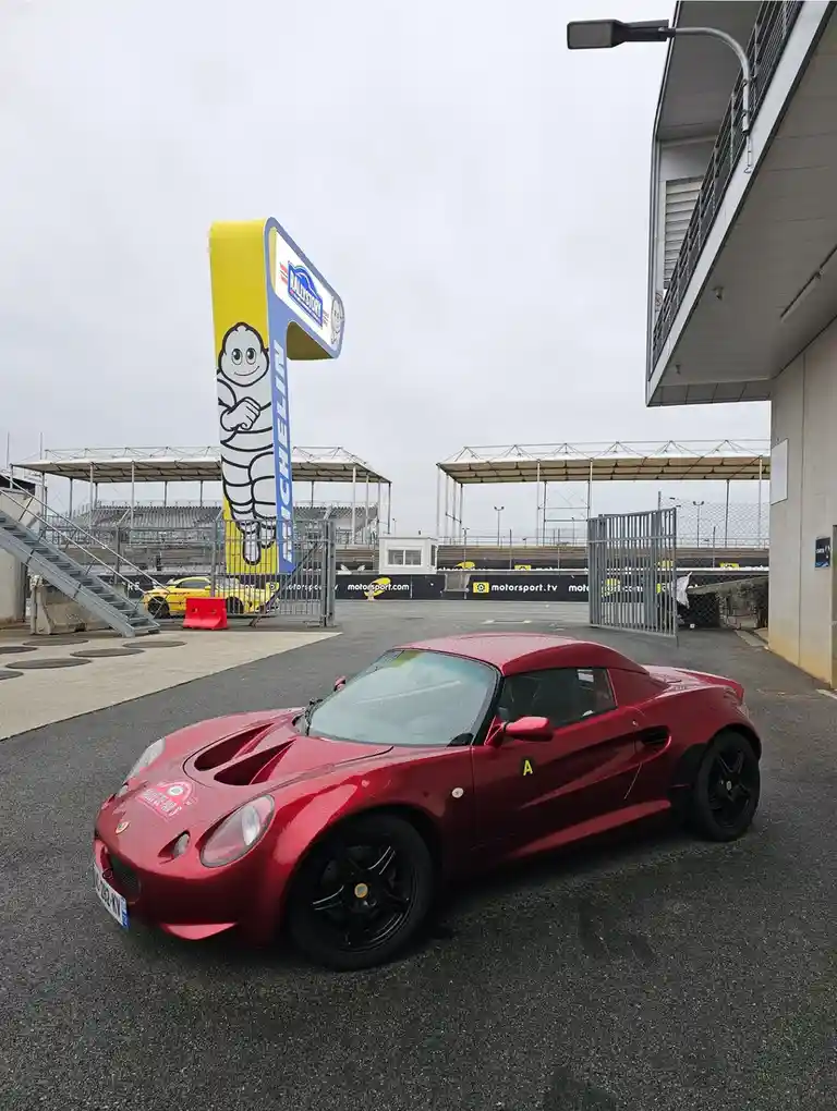 A red Lotus Elise sports car is parked in front of a large Michelin tire company sign featuring the Michelin Man mascot. The…