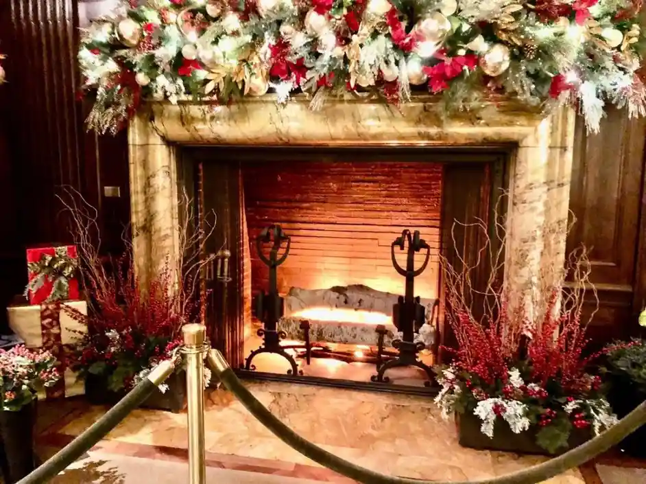 a lavishly decorated fireplace with a marble surround, adorned with red and gold holiday garlands, pinecones, and ornaments…