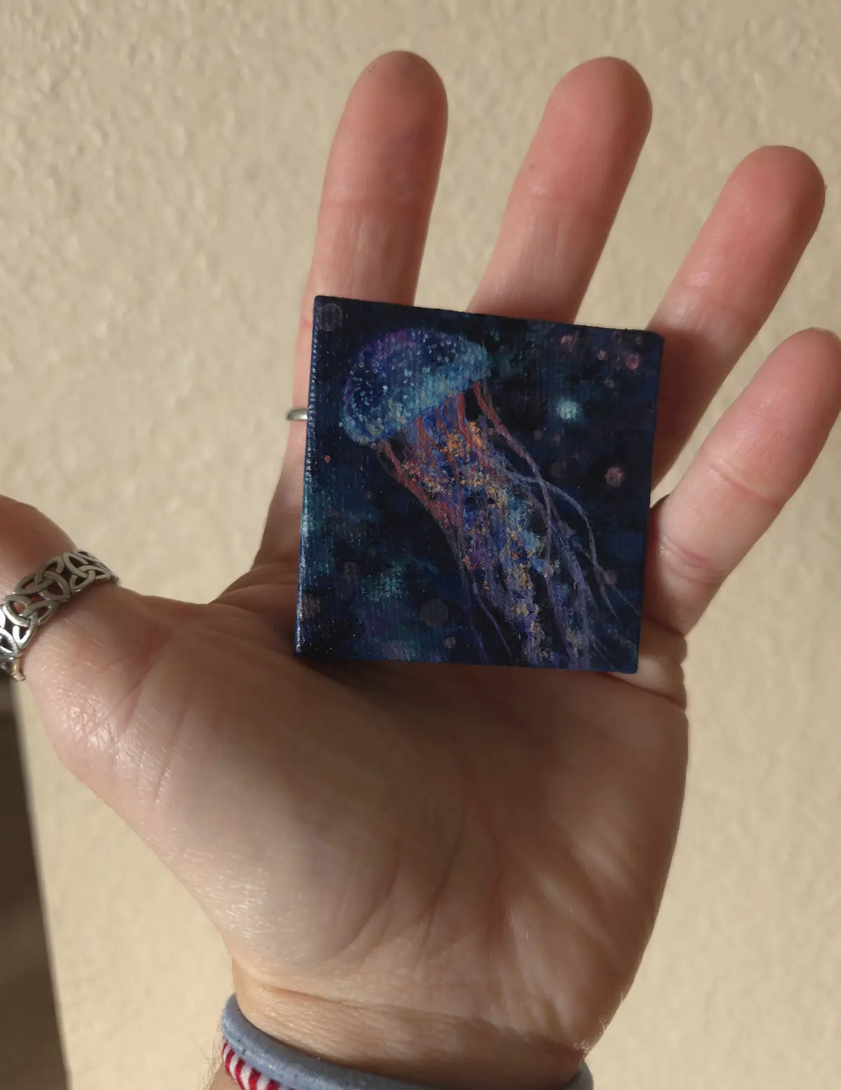 A hand holds a small square painting of a colorful jellyfish against a dark blue background with speckles. The hand has a…