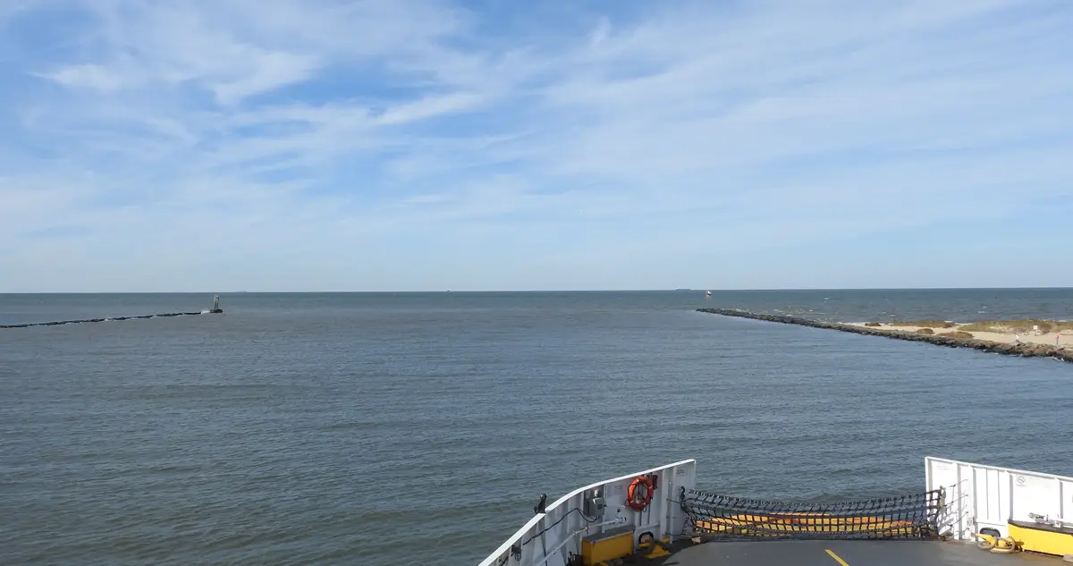 A photo taken from the back of a ferry shows a wide expanse of ocean under a partly cloudy sky, with a rocky jetty on the…