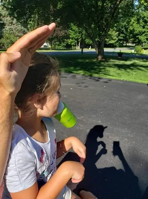 A young girl is drinking from a bright green sippy cup while an adult's hand is raised in a salute above her head. The…