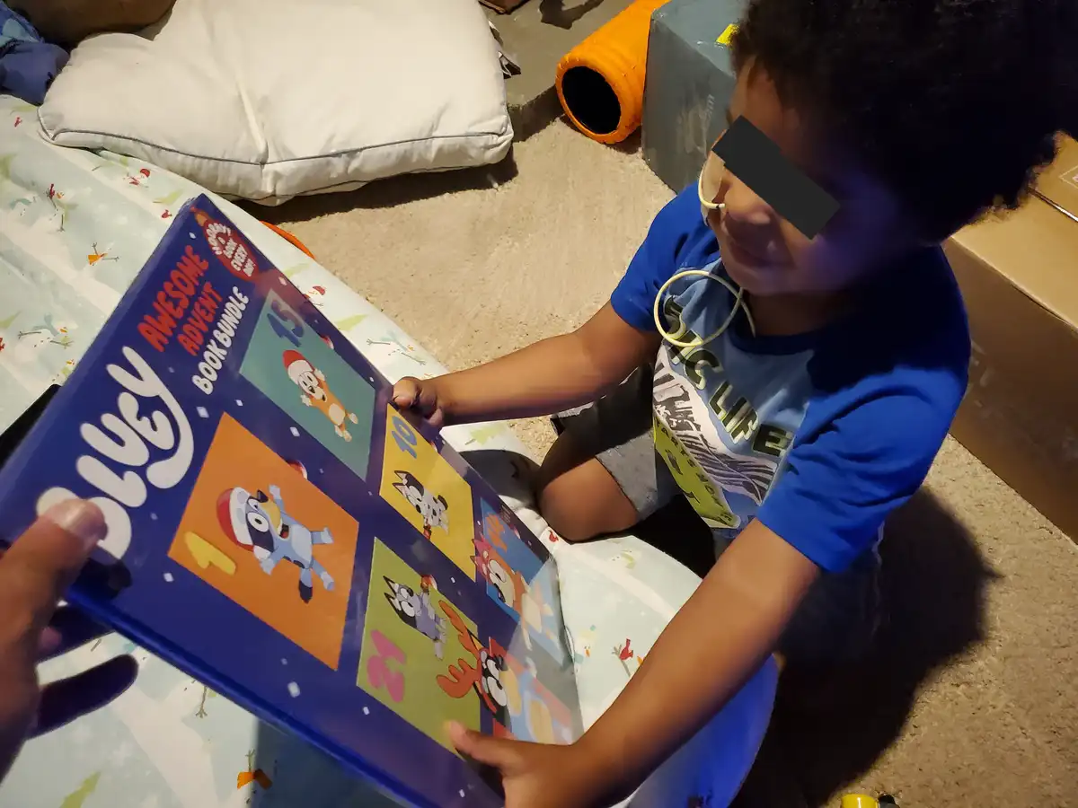 A young child with glasses and a blue shirt sits on a patterned blanket, looking at a 'Bluey Awesome Advent Book Bundle'…