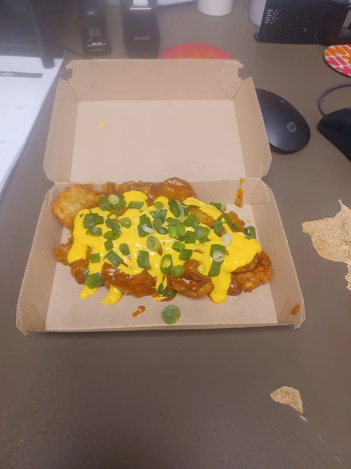A cardboard takeout box on a desk contains fried chicken pieces covered in orange sauce, melted yellow cheese, and chopped…