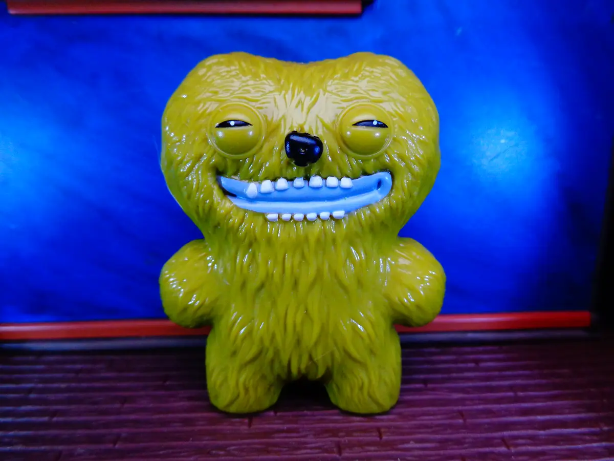 A yellow, furry, toy-like monster with a wide, toothy grin and half-closed eyes stands against a blue background. The…