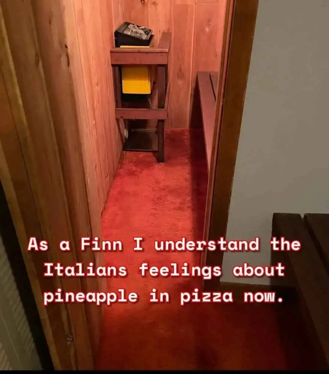 A narrow hallway with red carpet and wood-paneled walls leads to a small room with a wooden table holding a yellow trash can…