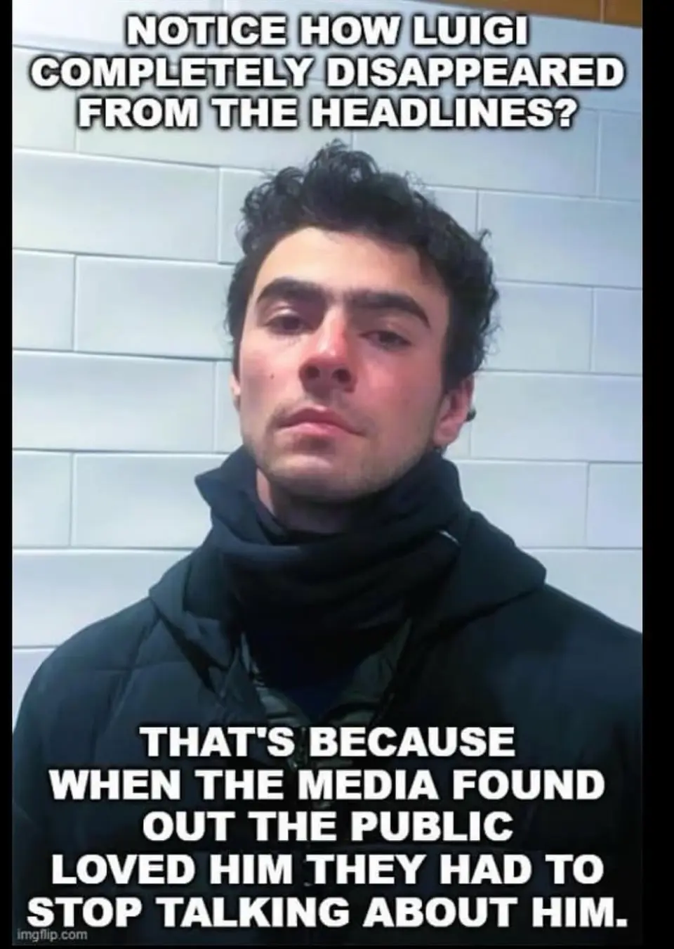 A man with dark curly hair and a black hooded jacket stands against a white tiled wall, looking directly at the camera with…