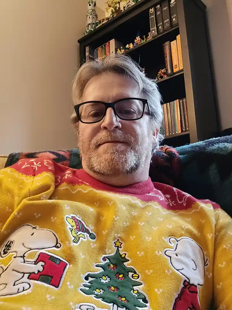 A man with glasses and a graying beard wears a festive yellow Christmas sweater featuring Snoopy, a Christmas tree, and…