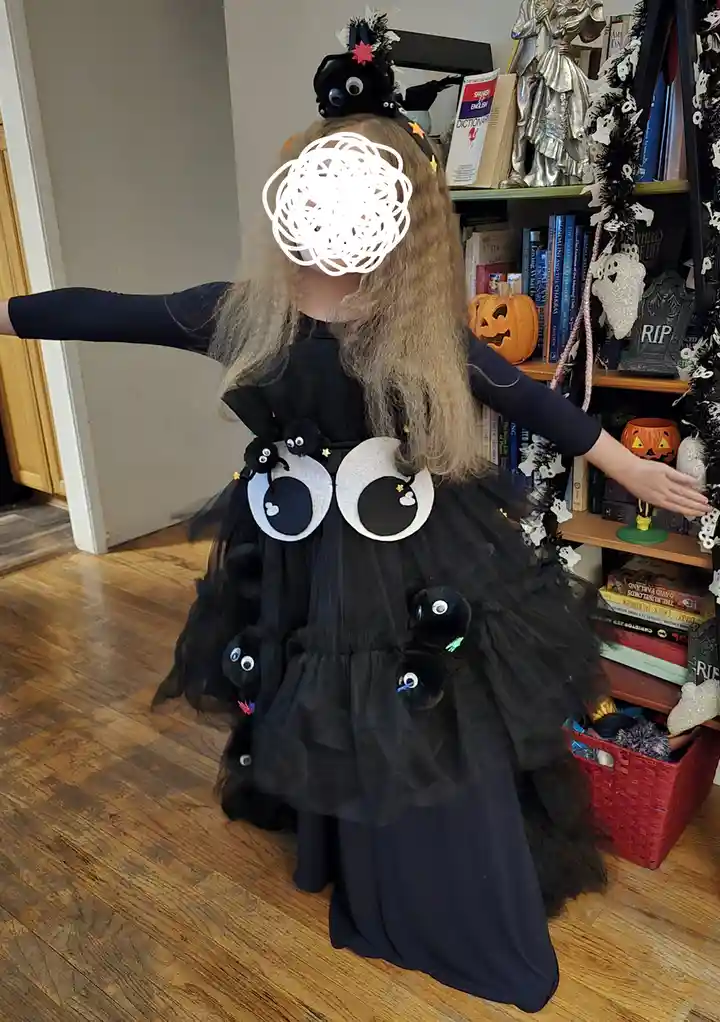 A child in a black Halloween costume with large googly eyes and spider decorations on the dress and headband poses with arms…