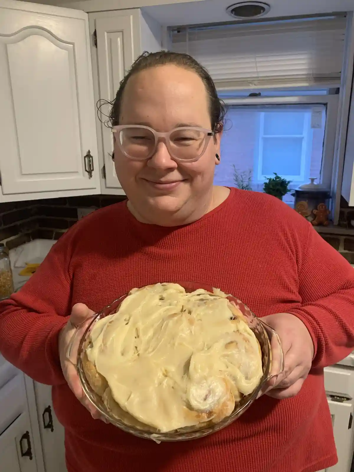 A person in a red sweater stands in a kitchen holding a glass dish filled with cinnamon rolls covered in a thick, creamy…