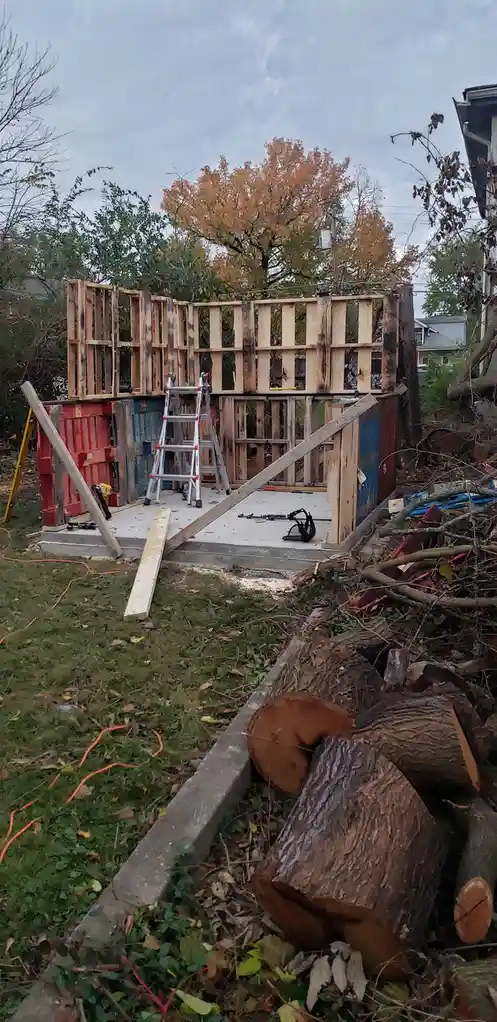 An outdoor construction site shows a partially built wooden structure on a concrete foundation, surrounded by tools…