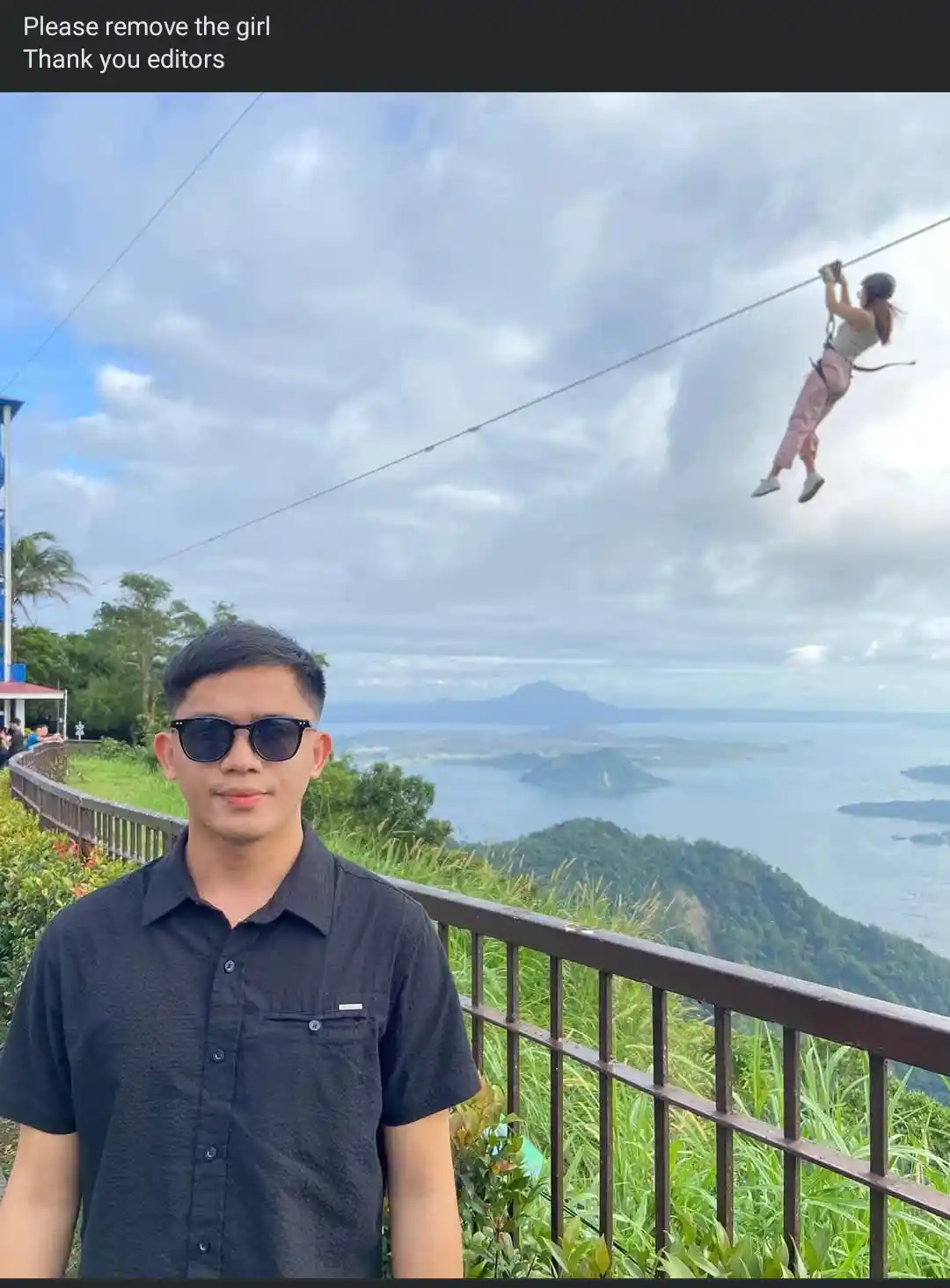 A man in a black shirt and sunglasses stands in the foreground on a scenic overlook with a railing, smiling at the camera…