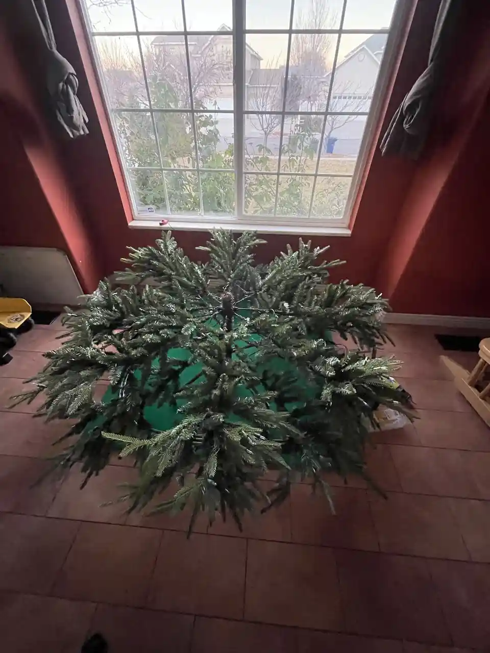 A flat, un-assembled artificial Christmas tree lies spread out on a tiled floor in front of a large window, its branches…
