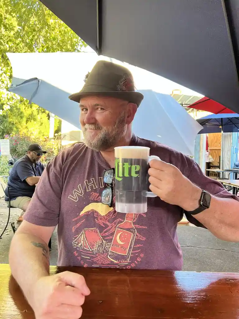 A middle-aged man with a beard and a brown fedora sits at an outdoor table under a large umbrella, holding up a large mug of…