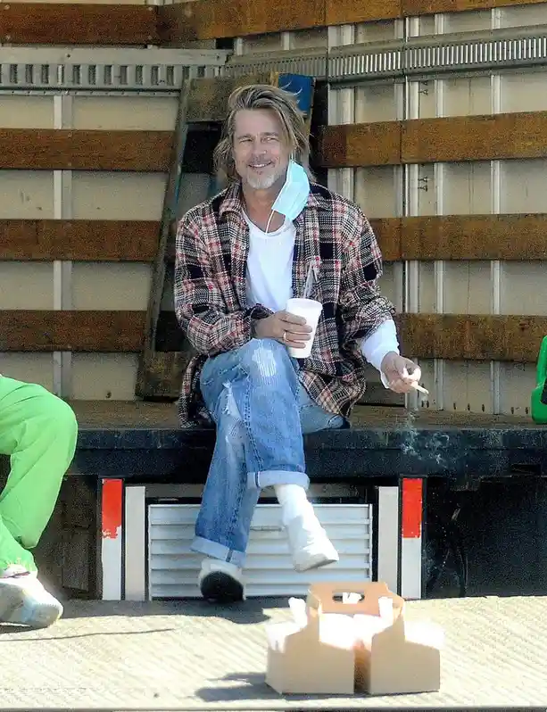 Brad Pitt is seen sitting casually on the tailgate of a truck, wearing a plaid shirt over a white t-shirt, ripped jeans, and…