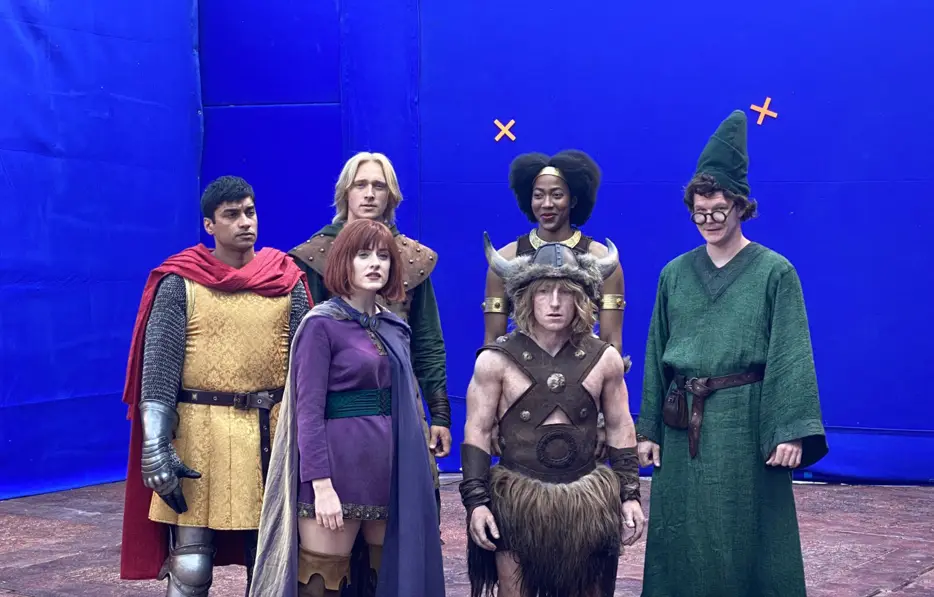 Five actors in fantasy costumes stand before a blue screen, with orange 'X' marks above the heads of the two actors on the…