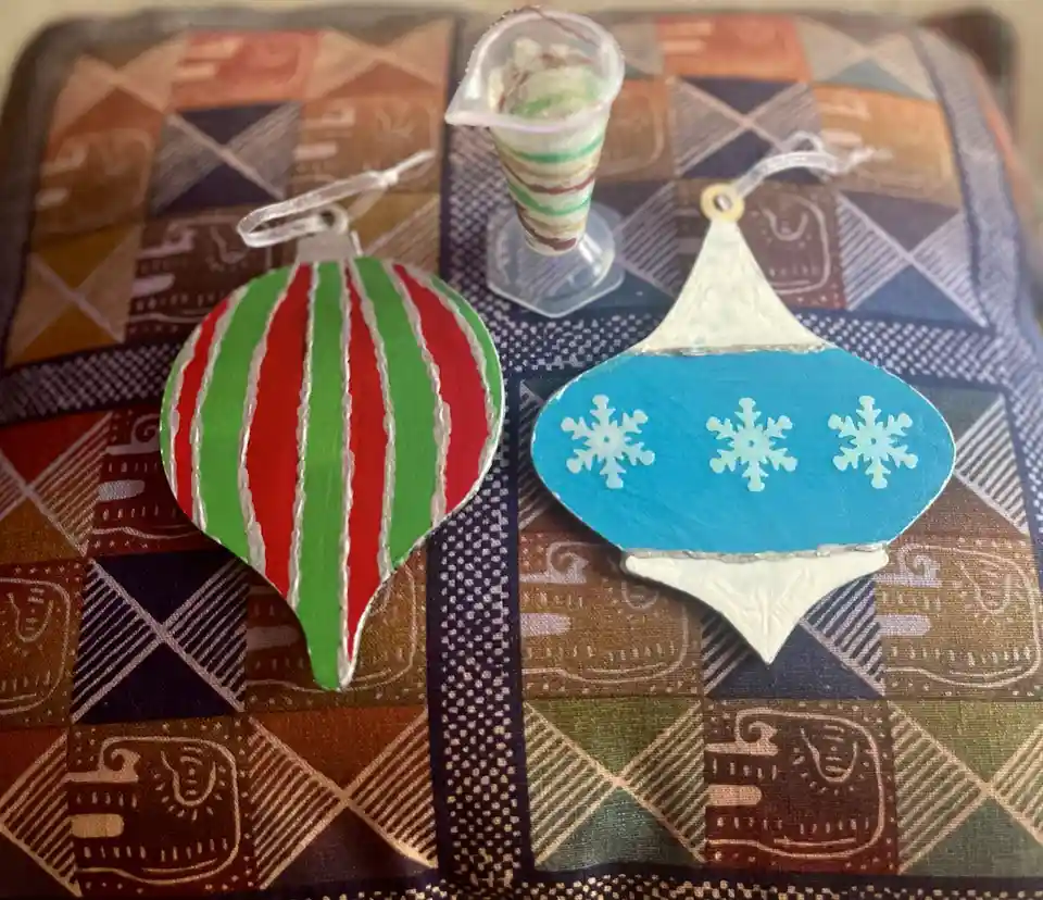 Two hand-painted Christmas ornaments sit on a patterned fabric surface, one striped red and green with white borders, the…