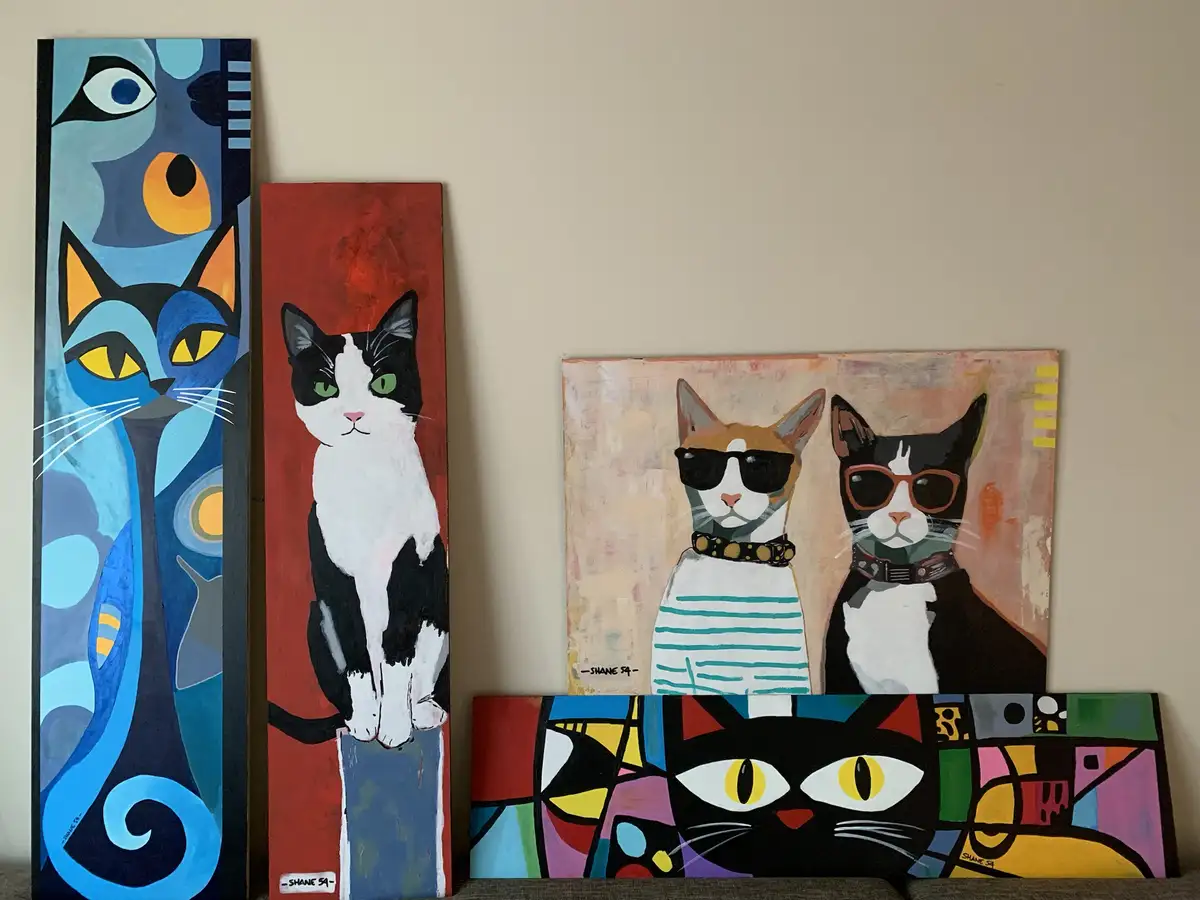 Four colorful, stylized paintings of cats are displayed leaning against a wall. The leftmost is a tall blue abstract cat…