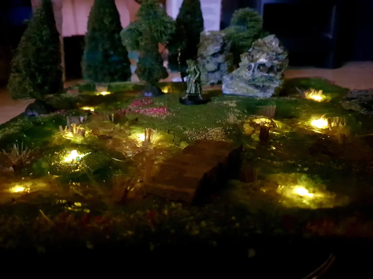A dimly lit tabletop miniature scene featuring a fantasy landscape with green mossy terrain, small trees, rocks, and a…